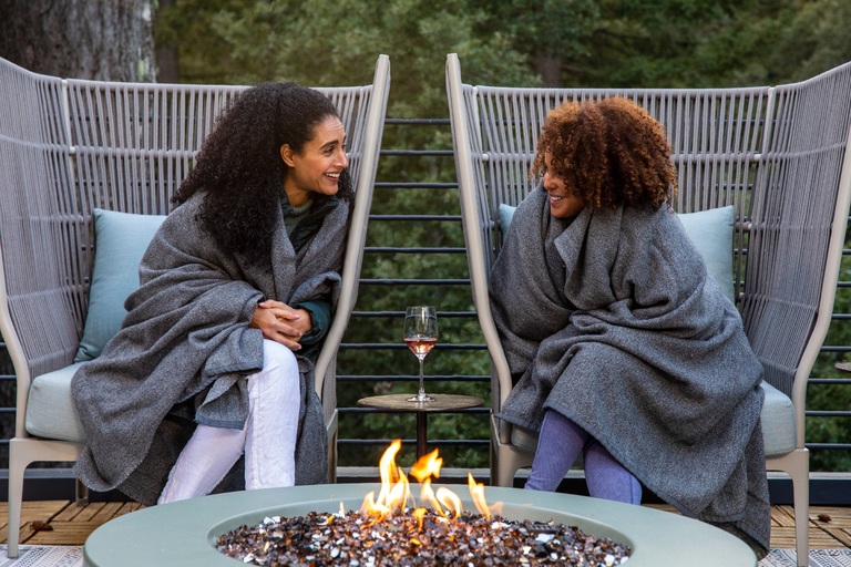 Two women cozy in blankets smile over a glass of wine