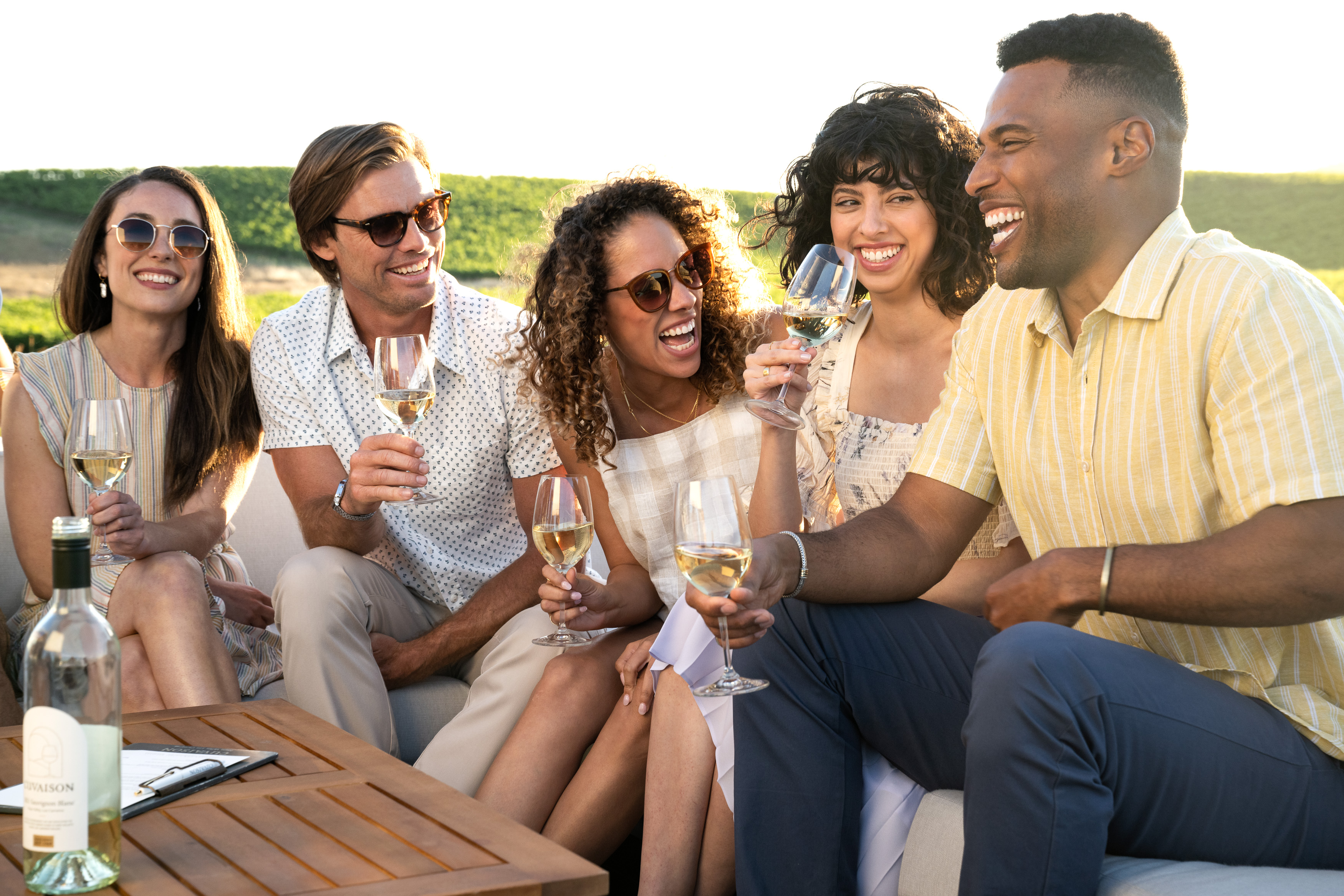 Group of people seated on the patio smiling while the sun sets over the vineyard behind them