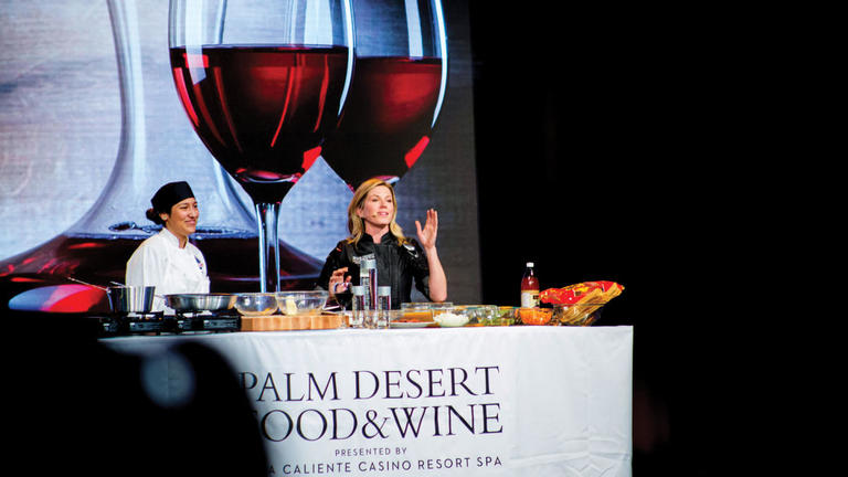 Two chefs presenting a cooking demonstration before a backdrop showing two large glasses of red wine.