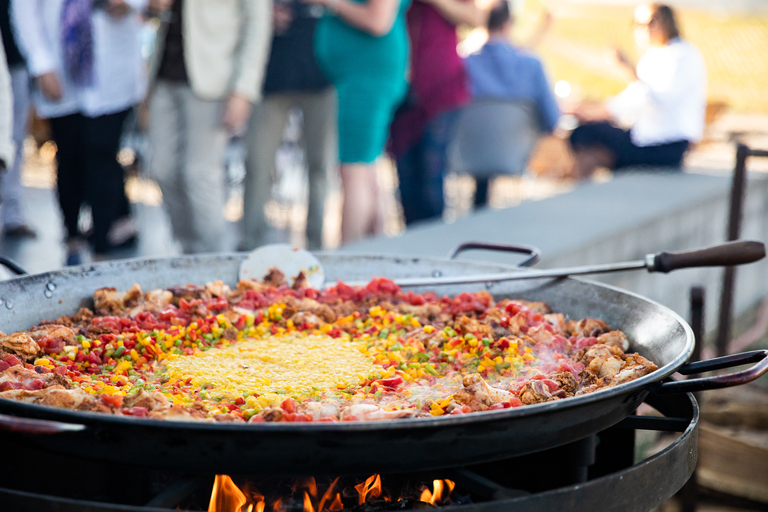 A large dish of Paella by a vineyard, with guests in the background